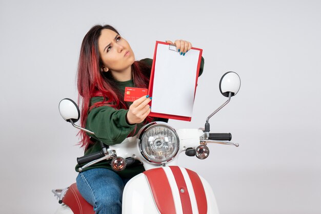 白い壁にノートと銀行カードを保持しているオートバイの正面図若い女性