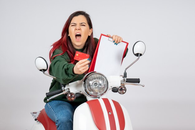 Вид спереди молодая женщина на мотоцикле, держащая банкноту и банковскую карту на белой стене