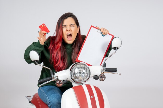 Front view young female on motorcycle holding note and bank card on white wall