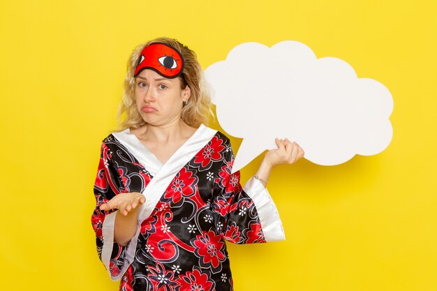 Front view young female in night robe eye mask holding huge white sign and posing on yellow wall sleep night bed model color