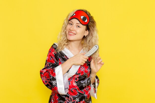 Front view young female in night robe and wearing eye mask brushing her hair on yellow wall sleep girl night bed model