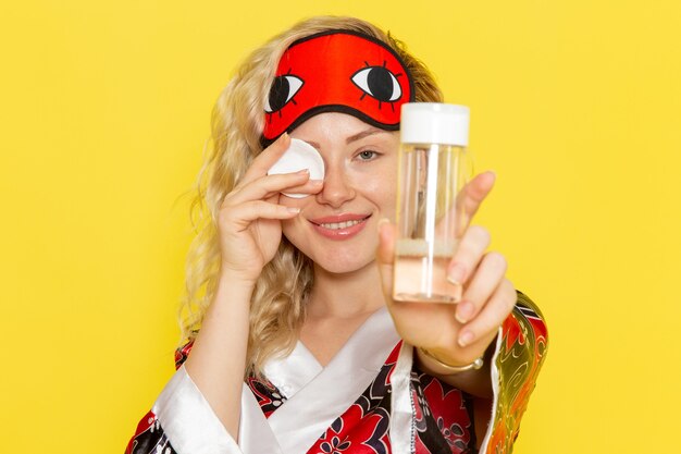 Front view young female in night robe and wearing eye mask cleaning her face from make-up smiling on yellow desk sleep girl night bed model