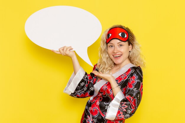 Front view young female in night robe and wearing eye mask holding big white sign on yellow desk sleep girl night bed model