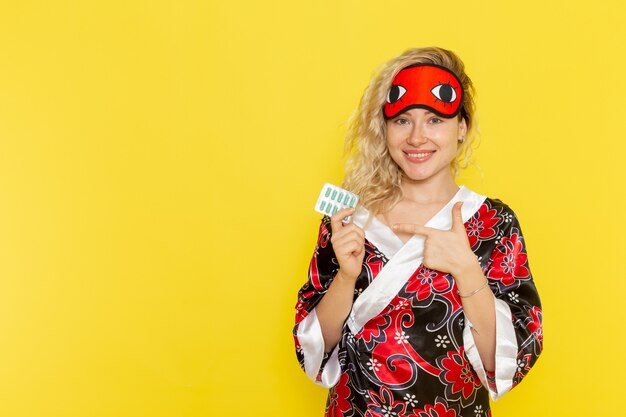 Front view young female in night robe and wearing eye mask preparing to sleep holding pills on the yellow wall sleep female darkness color night