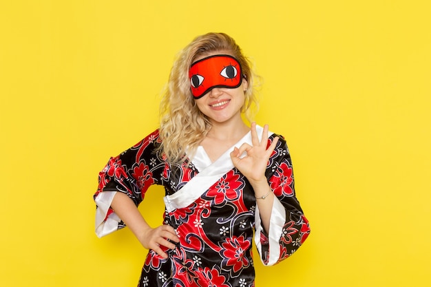Free photo front view young female in night robe and wearing eye mask preparing to sleep posing and smiling on yellow wall sleep female darkness color night