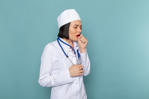 A front view young female nurse in white medical suit and blue stethoscope coughing