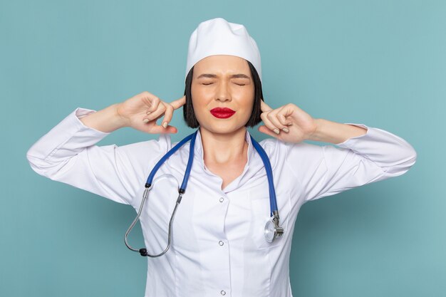 A front view young female nurse in white medical suit and blue stethoscope covering her ears