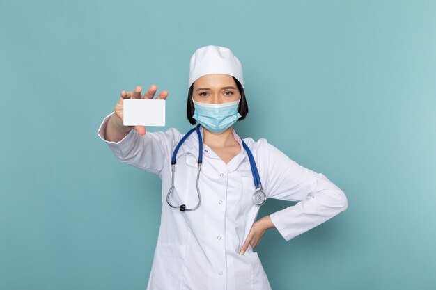 A front view young female nurse in white medical suit and blue stethoscope holding card