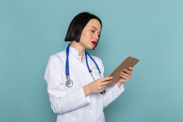 A front view young female nurse in white medical suit and blue stethoscope holding notepad on the blue desk medicine hospital doctor