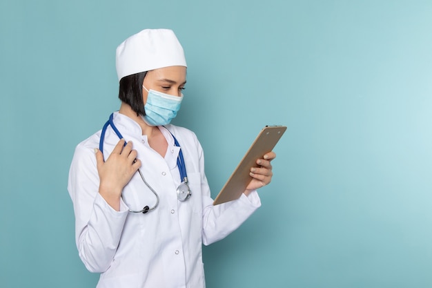 Вид спереди молодая медсестра в белом медицинском костюме и синем стетоскопе, держащая блокнот на синем столе врача больницы