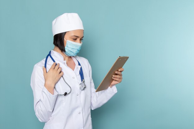 A front view young female nurse in white medical suit blue stethoscope holding notepad on the blue desk medicine hospital doctor