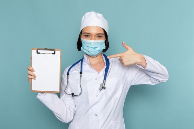 A front view young female nurse in white medical suit and blue stethoscope holding notepad in mask