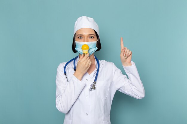 A front view young female nurse in white medical suit and blue stethoscope and mask