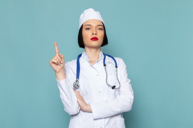파란색 책상 의학 병원 의사에 제기 손가락으로 포즈 흰색 의료 양복과 파란색 청진기에 전면보기 젊은 여성 간호사