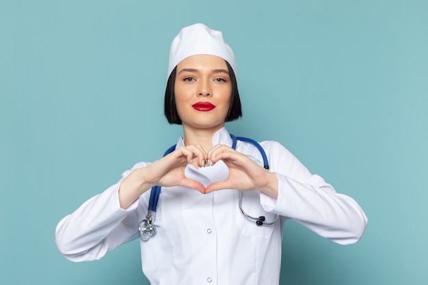 Una giovane infermiera femminile di vista frontale in vestito medico bianco e stetoscopio blu che mostra il segno del cuore sul medico blu dell'ospedale della medicina dello scrittorio