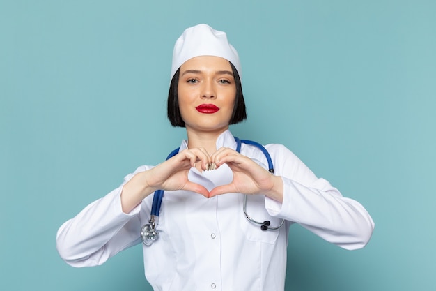 A front view young female nurse in white medical suit and blue stethoscope showing heart sign on the blue desk medicine hospital doctor