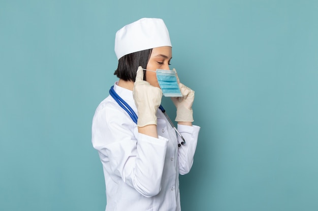 Вид спереди молодая медсестра в белом медицинском костюме и синем стетоскопе в маске на синем столе врача больницы