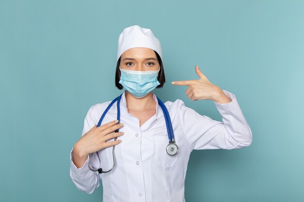 A front view young female nurse in white medical suit and blue stethoscope wearing a mask on the blue desk medicine hospital doctor