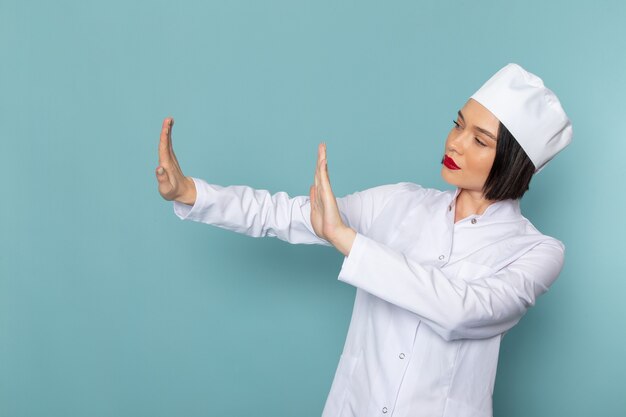 Una giovane infermiera femminile di vista frontale in vestito medico bianco avverte con il segno delle mani sul medico dell'ospedale della medicina dello scrittorio blu