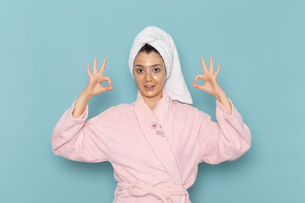 青い壁のクリーニング美容きれいな水セルフケアクリームシャワーのシャワーの後のピンクのバスローブの正面図若い女性