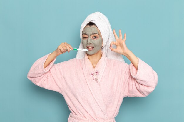 Front view young female in pink bathrobe after shower cleaning her teeth on light-blue wall beauty water selfcare shower clean