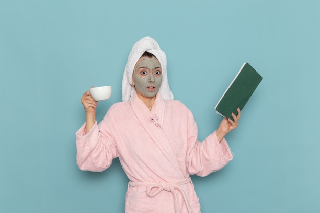 Вид спереди молодая женщина в розовом халате после душа, пьющая кофе на синей стене, чистка красоты, чистая вода, крем для ухода за собой, душ
