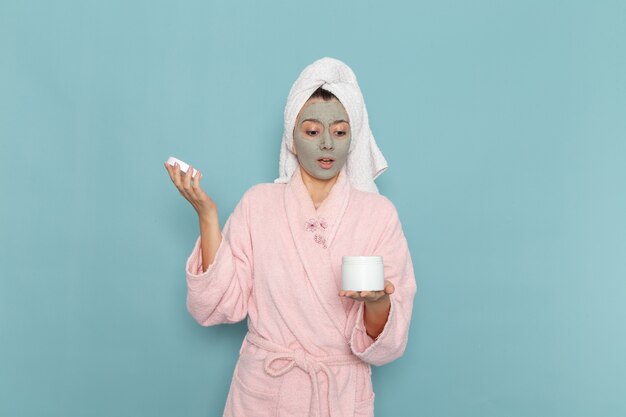 Вид спереди молодая женщина в розовом халате после душа, держащая крем на синей стене, косметическая водяная ванна, крем для душа, ванная комната