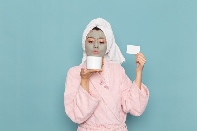 Front view young female in pink bathrobe after shower holding cream and card on blue wall beauty water cream selfcare shower bathroom