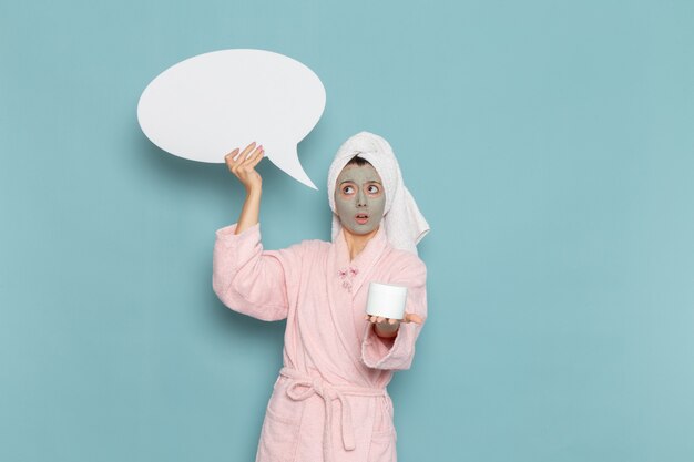 Front view young female in pink bathrobe after shower holding cream and sign on blue wall beauty water cream selfcare shower bathroom