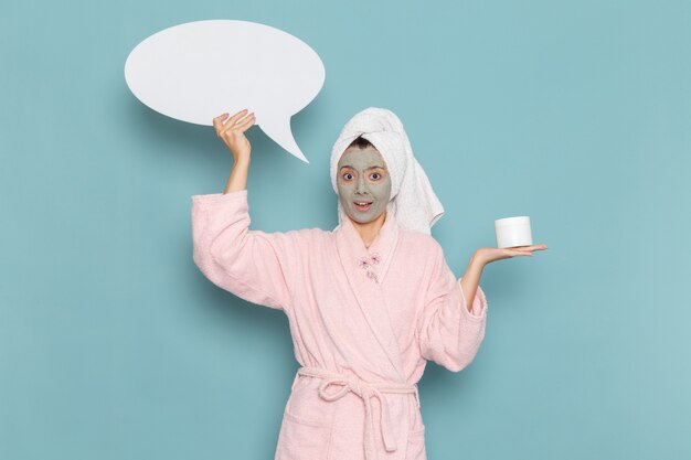 Front view young female in pink bathrobe after shower holding huge white sign on blue desk beauty water cream selfcare shower bathroom