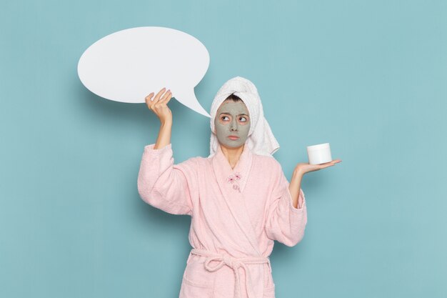 Front view young female in pink bathrobe after shower holding huge white sign on blue wall beauty water cream selfcare shower bathroom