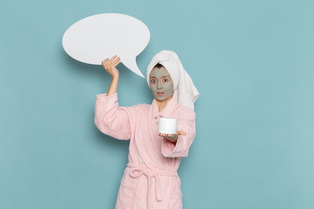 Front view young female in pink bathrobe after shower holding huge white sign on light blue wall beauty water cream selfcare shower bathroom