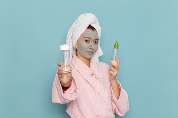 Front view young female in pink bathrobe after shower holding sprays on the light blue wall beauty water cream selfcare shower bathroom