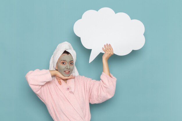 Front view young female in pink bathrobe after shower holding white sign on light-blue wall beauty water selfcare shower clean