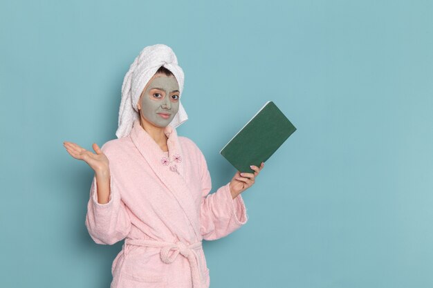 Front view young female in pink bathrobe after shower reading copybook on the blue wall beauty water bath cream selfcare shower bathroom