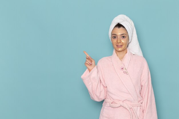 Вид спереди молодая женщина в розовом халате после душа, улыбающаяся на синей стене, косметический крем с водой, самообслуживание, душ, ванная комната