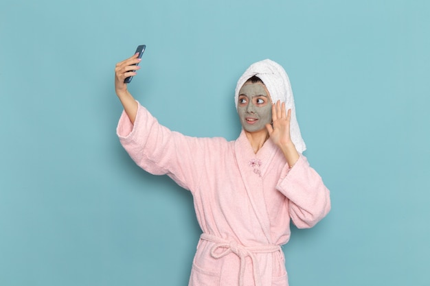 Front view young female in pink bathrobe after shower taking a selfie on blue wall beauty water cream selfcare shower