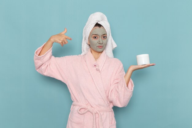 青い壁にクリームを保持している彼女の顔にマスクを付けたシャワーの後のピンクのバスローブを着た若い女性の正面図シャワークリーニング美容セルフケアクリーム