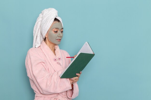 Вид спереди молодая женщина в розовом халате после душа пишет на синей стене косметический водный крем для душа в ванной комнате