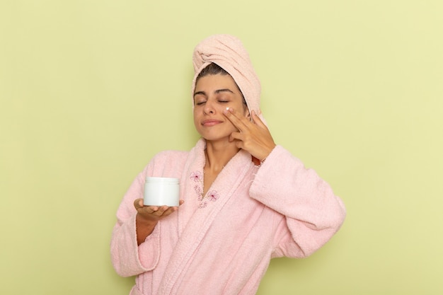 Front view young female in pink bathrobe applying face cream on a green surface