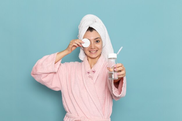 Front view young female in pink bathrobe cleaning her face from make-up on blue floor cleaning beauty selfcare cream shower