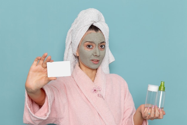 Front view young female in pink bathrobe holding card and sprays on the blue wall beauty water bath cream selfcare shower