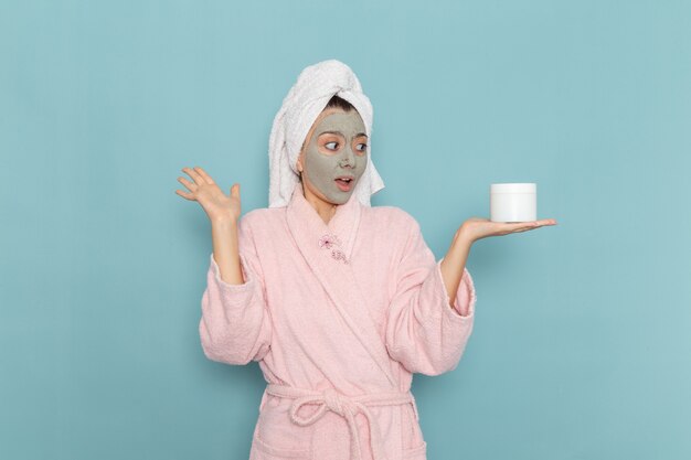 Front view young female in pink bathrobe holding cream for face on blue wall shower cleaning beauty self-care cream