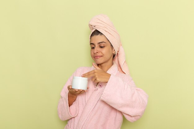 Front view young female in pink bathrobe holding cream on a green surface