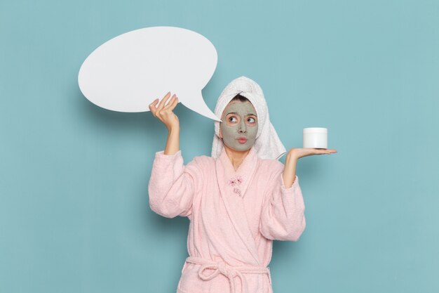 Front view young female in pink bathrobe holding huge white sign on blue wall shower cleaning beauty self-care cream