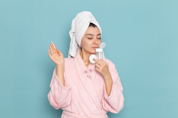 Вид спереди молодая женщина в розовом халате, держащая средство для снятия макияжа на синей стене, чистка красоты чистой водой, крем для ухода за собой, душ