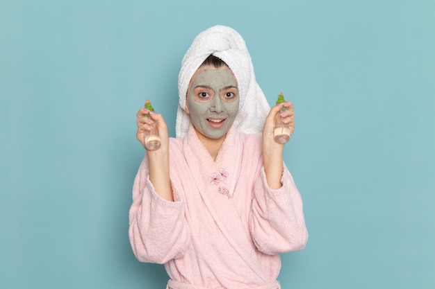Front view young female in pink bathrobe holding sprays on blue wall shower cleaning beauty self-care cream