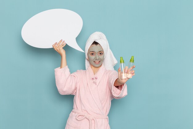 Front view young female in pink bathrobe holding white sign and sprays on the blue wall beauty water bath cream selfcare shower