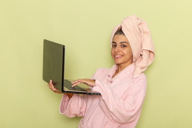 Вид спереди молодая женщина в розовом халате с помощью ноутбука на зеленой поверхности