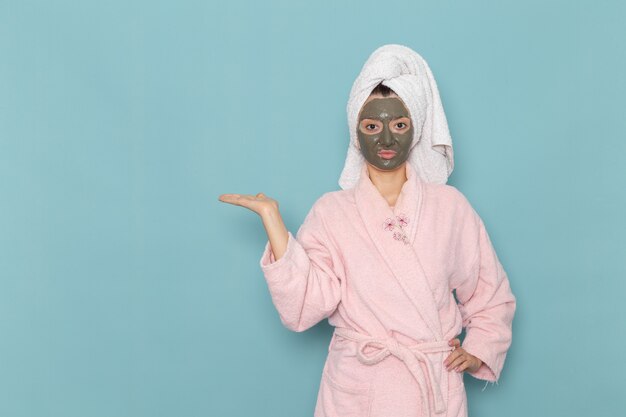 파란색 벽 샤워 청소 아름다움 셀프 케어 크림에 그녀의 얼굴에 마스크와 핑크 목욕 가운에 전면보기 젊은 여성